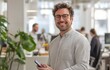 © Georgii - Confident young professional man smiling in a modern office environment while holding a smartphone, showcasing casual work style and technology. Ideal for business and technology-related themes.
