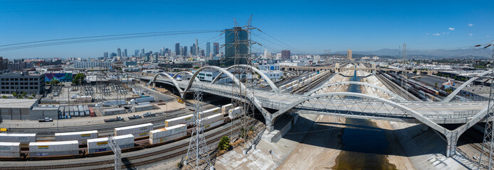 The image captures the Los Angeles River's concrete channel with the Sixth Street Viaduct. Downtown skyscrapers and freight trains add to the urban scene.