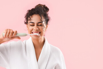 Poster - Young African-American woman in bathrobe brushing teeth on pink background