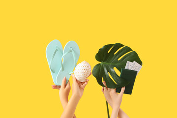 Canvas Print - Female hands with flip-flops, shell, passport with tickets and monstera leaf on yellow background. Travel concept