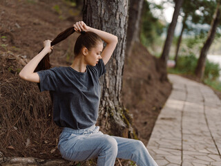 Young woman sitting on path in nature, tying her long hair back, wearing casual grey t shirt and light trousers, focused and serene expression Natural lifestyle concept