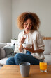 © Daniel - Vertical portrait of young multiracial woman eating breakfast sitting on sofa at home living room.