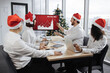 © sofiko14 - Happy team of diverse business people in Santa hats listening their middle aged male bearded chief, showing dynamics of r world market, using digital TV screen at office decorated with Christmas tree.