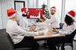 © sofiko14 - Happy team of diverse business people in Santa hats listening their middle aged male bearded chief, showing dynamics of r world market, using digital TV screen at office decorated with Christmas tree.