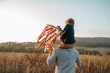 © Halfpoint - Rear view of father walking with daughter on his shoulders across autumn meadow. Girl holding wreath with ribbons.