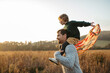 © Halfpoint - Father walking with daughter on his shoulders across autumn meadow. Girl holding wreath with ribbons.