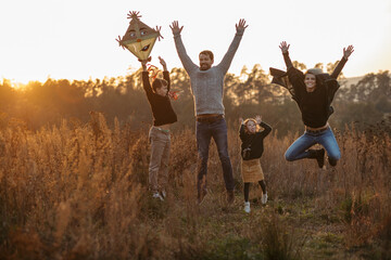 Wall Mural - Portrait of young family flying a kite in the autumn nature, jumping in the middle meadow during sunset. Indian summer.