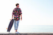 © Raul Mellado - back view of young hipster male skater in trendy summer outfit carrying skateboard while standing on embankment against sea and cloudless sky in summer day