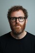 © video rost - A close-up portrait of a man with curly hair and a red beard, wearing black glasses and a serious expression. The soft lighting and neutral gray background create a calm and composed atmosphere