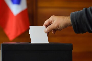 A significant act of casting a vote is captured as a hand places a ballot in a box, with a national flag draped in the backdrop, reflecting civic engagement.