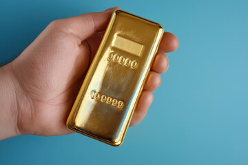 A close-up of a hand holding a gold bar against a blue background. The gold bar has embossed alphanumeric characters.