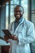 © Georgii - Confident and happy doctor in a white coat with glasses holding a tablet, smiling in a modern medical office setting