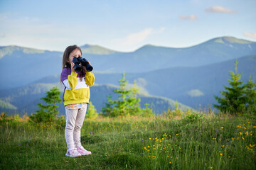 Wall Mural - Young girl stands in grassy meadow, looking through binoculars, wearing yellow and purple sweatshirt. Behind her, rolling green hills and mountains stretch into distance under clear blue sky.