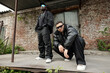© pressmaster - Two men standing and squatting near outdoor building entrance, wearing casual streetwear and sunglasses. Brick wall background and ivy climbing up on side, creating urban vibe