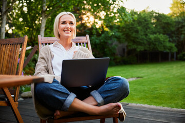 Smiling mature woman using laptop in garden sitting on chair in backyard