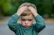 © Rohit k  - Stressed young boy outdoors, looking down with a frustrated expression, holding his head in worry. His green shirt highlights his troubled emotions