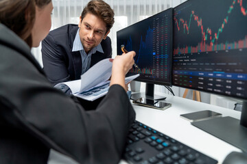 Two business traders discussing value business statistic graph company on paper report analyzing with dynamic stock exchange market currency real time website surround pc screens at office. Postulate.