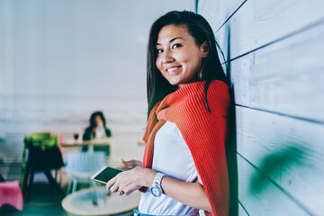 Half length portrait of cheerful asian female blogger with smartphone device in hand looking at camera during leisure time in publicity area near wall, concept of technology and communication