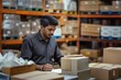 © Pete - Indian man works in shipping office. Man packing boxes in warehouse. Logistic dispatcher prepares records at desk. Man in black shirt focused on paperwork. Warehouse filled with shelves stacked with