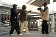 © pressmaster - Three individuals filming under an architectural structure holding professional camera. One person is holding camera while two others gesturing and engaging in discussion