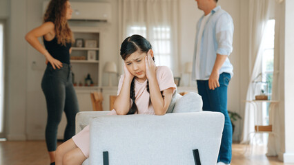 Annoyed and unhappy young girl sitting on sofa trapped in middle of tension by her parent argument in living room. Unhealthy domestic lifestyle and traumatic childhood develop to depression Synchronos