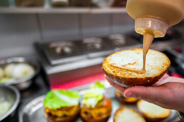 Wall Mural - Close-up of a hand applying sauce on a burger bun in a kitchen, with blurred burgers in the background. Perfect for food preparation, culinary arts, and fast food themes.