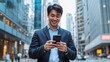 © Adi - Young happy Asian businessman executive, Japanese business man investor standing in big city street with downtown buildings using cell phone looking at smartphone checking financial apps on mobile