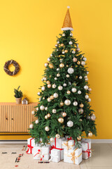 Canvas Print - Decorated Christmas tree with gift boxes, silver balls, Santa hat and wooden cabinet near yellow wall