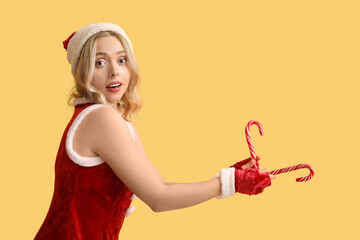 Canvas Print - Beautiful young woman dressed as Santa Claus with candy canes on yellow background