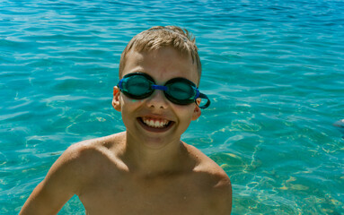 Happy boy wearing swimming goggles smiles while swimming in crystal clear turquoise water on a sunny summer day. Concept of childhood joy, outdoor activities, and water fun. High quality photo