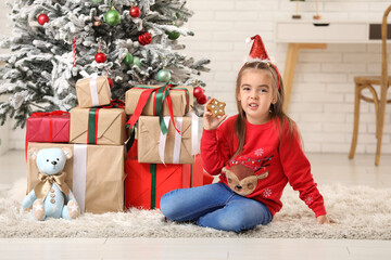 Wall Mural - Cute little girl in Santa hat headband with sweet cookie and gift boxes at home on Christmas eve