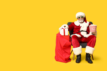 Wall Mural - Santa Claus holding bucket of tasty popcorn and bag with gifts on yellow background