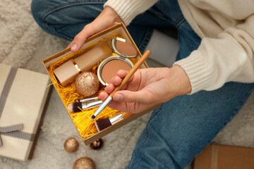 Poster - Woman holding Christmas gift boxes with makeup brushes and cosmetics at home, above view