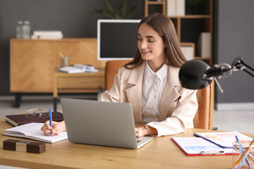 Canvas Print - Beautiful young happy businesswoman working with laptop while writing in notebook at table in office