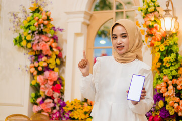 girl veil wearing a white dress muslim cheerful to the camera hand holding a mobile phone screen, smart phone, cellphone while pointing up in a luxurious interior. for fashion, religious content