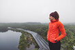 © VisualProduction - A smiling woman in an orange puffy jacket stands by a misty lake surrounded by greenery. She wears a headband and has a relaxed posture, enjoying the serene outdoor environment. The fog creates a tran