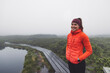 © VisualProduction - A smiling woman in an orange puffy jacket stands by a misty lake surrounded by greenery. She wears a headband and has a relaxed posture, enjoying the serene outdoor environment. The fog creates a tran