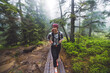 © VisualProduction - A person walking on a wooden path through a misty forest. The individual is wearing a gray long-sleeve shirt and black leggings, carrying an orange backpack. The surrounding trees are lush and green,