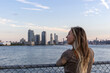© Pedro - A woman gazes at the New York City skyline during sunset by the river, capturing the vibrant colors and iconic buildings in the evening light