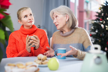 Mature mother and her adult daughter, who came to visit her for Christmas, drink tea and chat while sitting at a table in ..the room