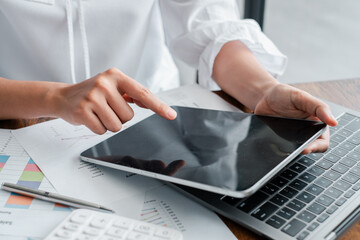 Close-up of a business professional using a tablet and laptop for data analysis, surrounded by charts and documents.