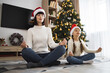 © sofiko14 - Caucasian mother and daughter sitting in yoga positions, wearing Santa hats, meditating together in cozy living room decorated with Christmas tree.
