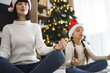 © sofiko14 - Caucasian mother and daughter sitting in yoga position wearing Santa hats meditating together by Christmas tree creating peaceful holiday atmosphere. Tree decorated with lights creates warm.