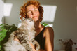 © larisikstefania - portrait red-haired curly young woman with pet gray fluffy cat in arms, lit by bright sun
