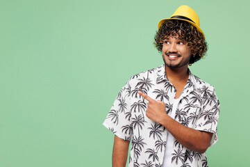 Wall Mural - Young happy fun smiling Indian man he wearing beach shirt hat rest near hotel pool point index finger aside on empty area isolated on plain green color background. Summer vacation sea sun tan concept.