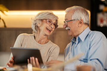 Cheerful senior couple laughing at dining table and holding tablet.