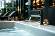 © Natalia - Modern metallic faucet with running water into a large bathtub in a luxurious spa or bathroom setting with blurred background featuring lounge chairs and warm lighting.