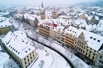 Canvas Print - Aerial view of a historic European city covered in snow, with prominent buildings, a church, and a curved street lined with trees and snow-covered rooftops.