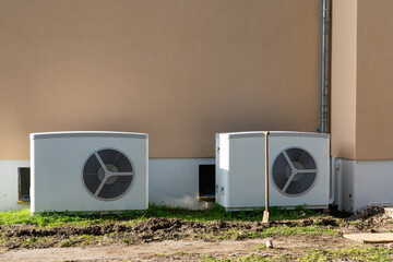 Two air conditioning units are placed next to a residential building, positioned on a grass patch. The sun casts shadows, indicating a clear, bright day.