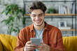 © Nadezda Ledyaeva - Young man holding his mobile phone in his hands as he relaxes at home on a comfortable sofa looking at the camera with a smile